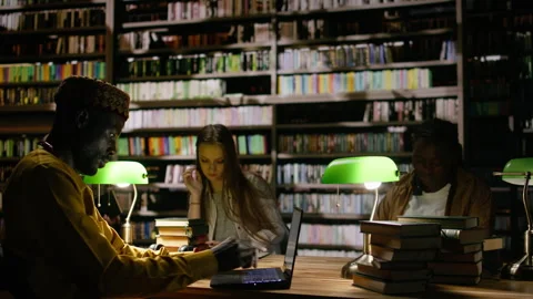 Side profile of black adult learner typing on laptop in dark library study room Stock Footage 337230123