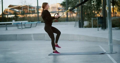 Side shot through metal net fence of young woman hard workout using TRX loops Stock Footage 123662890