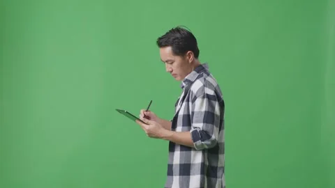 Side View Of Asian Man Taking Note On Tablet While Walking On Green Screen Stock Footage 248007845