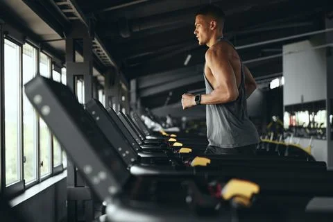 Side view of athletic man running on treadmill preforming routine exercise in Stock Photos