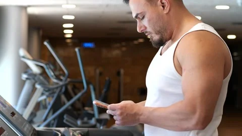 Side view of an athletic man using his smartphone while resting on a treadm.. Video stock 344048936