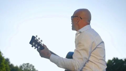 Side view of a bald man plays guitar and sings in park. Guitarist touching Stock Footage 112828818