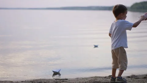 Side view of boy standing on sandy shore with paper boats. Child looks across Stock Footage 336382348