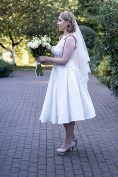 Side view of a bride in a tea-length white wedding dress and lace veil, holding  Stock Photos