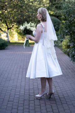 Side view of a bride in a tea-length white wedding dress and lace veil, holding  Stock Photos