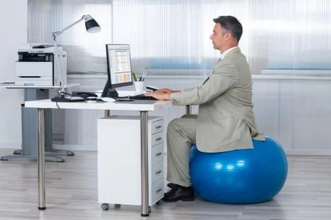 Side view of businessman using computer while sitting on exercise ball at off Stock Photos