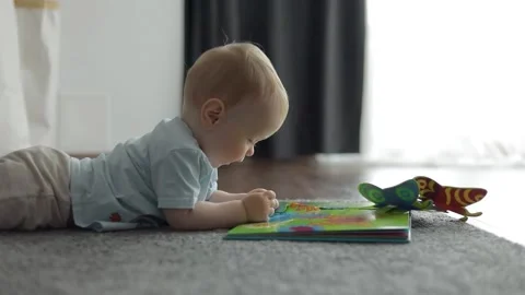 Side view. Cheerful baby lying on the carpet and reading a book Stock Footage 133440960
