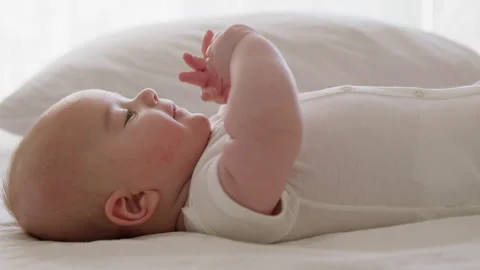 Side view, close-up, baby in white sleeveless bodysuit, lying on the bed on his Video stock 304695730