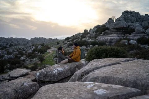 Side view of couple talking while sitting on rock against sky at sunset Stock Photos