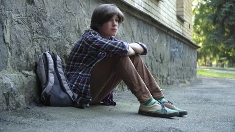 Side view of crying bullied schoolboy sitting outdoors at rock wall. Wide shot Video stock 143387301