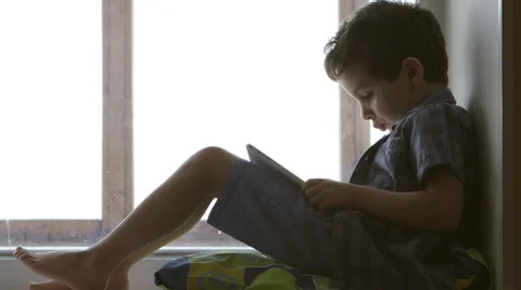 SIDE VIEW: A cute little boy uses a white tablet PC on a windowsill at home Stock Footage 66190664