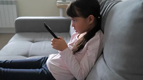 Side view of cute little kid sitting on couch watches TV with interest, holding Video stock 219393086