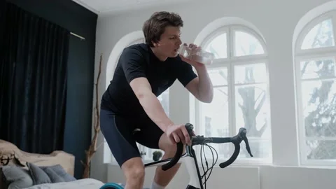 Side view of cyclist training on an exercise bike and drinking water at home 스톡 동영상 171117452