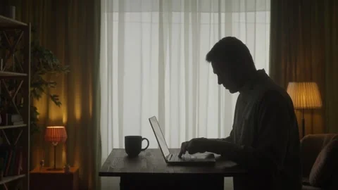 Side view of a dark silhouette of a man at a table in front of a laptop. A man Stock Footage 245490471