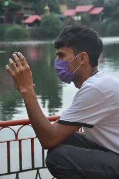 Side view of a guy using mobile phone, wearing face mask sitting beside the lake Stock Photos