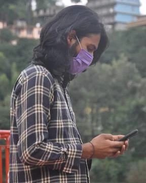Side view of a Indian long haired young man wearing face mask with using phone  Stock Photos