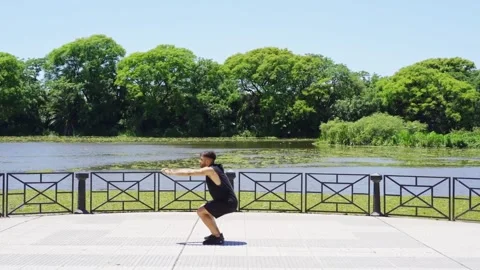 Side view of a  Latino man doing squats by the lakeside in a city park Video stock 300104675