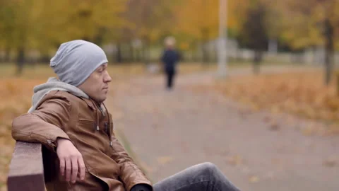 Side view of a man in a gray hat sits and waits on a bench in a city park Stock Footage 163562250