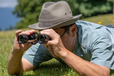 A side-view of a man lying down on a grass field and using binoculars. Stock Photos