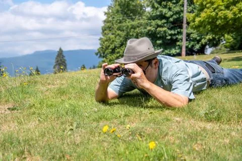 A side-view of a man lying down on a grass field and using binoculars. Stock Photos