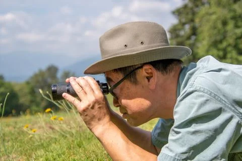 A side-view of a man lying down on a grass field and using binoculars. Stock Photos