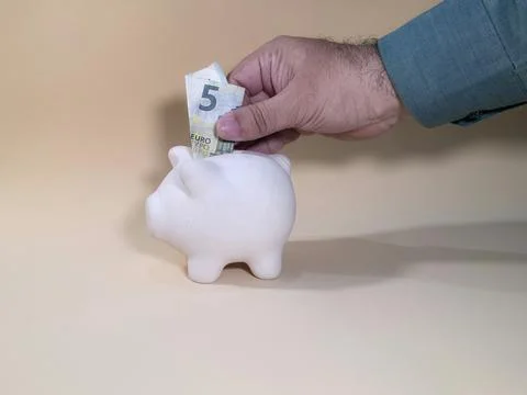 Side view of a man's hand depositing a 5 Euro note into a white piggy bank Foto stock