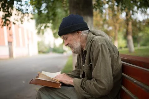 Side view of old homeless man with long grey hair reading a book. Stock Photos