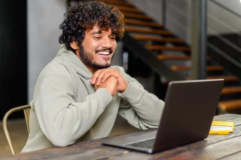 Side view at optimistic Indian man wearing hoodie sitting at desk and using Stock Photos