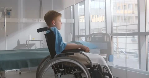 Side view of pensive kid sitting on wheelchair looking out of window in hospital Stock Footage 154123185