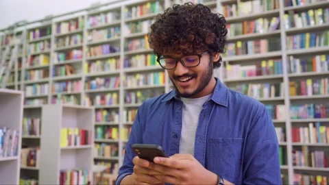 Side view portrait of happy young Indian student chatting by phone in library Stock Footage 164931744