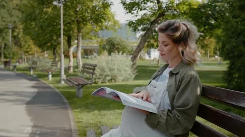 Side view of pregnant sitting on a bench reading book. Video stock 300018602
