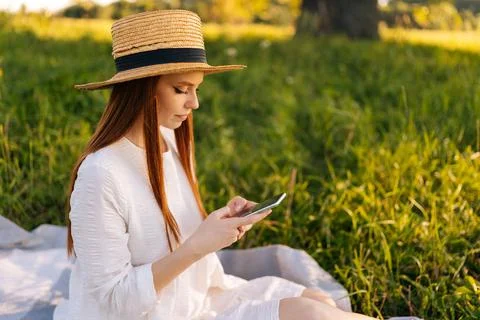 Side view of pretty redhead young woman in straw hat and white dress using Stock-Fotos
