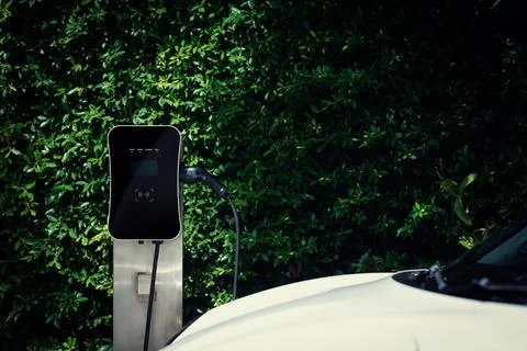 Side view progressive EV car with charging station, green foliage background. Stock Photos