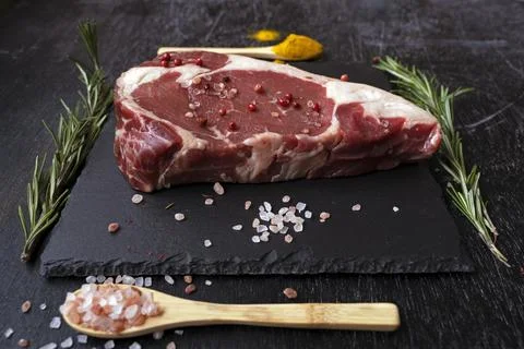 Side view raw rib eye steak on a square black slate board with two spoons and Stock Photos