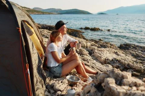 Side view of romantic couple relaxing near tent at rock beach. Stock Photos