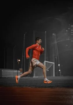 Side view of a runner on a red track with goal posts behind at night Stock Photos