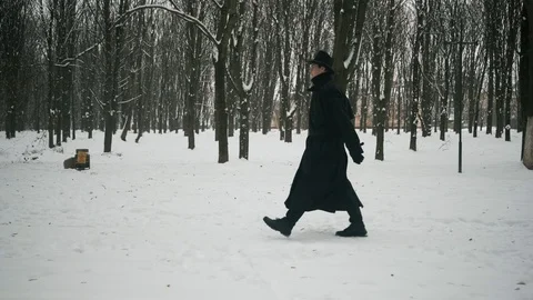 Side view shot: A man in a black cloak and hat walks in the winter park. 60fps Stockbeeldmateriaal 102195872