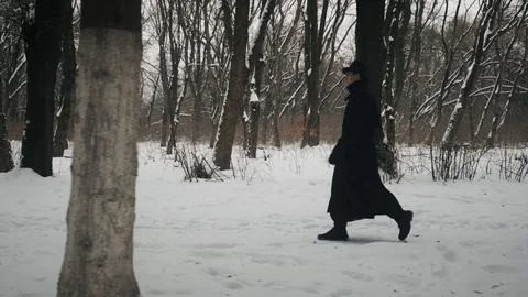 Side view shot: A man in a black cloak and hat walks in the winter park. 60fps Stockbeeldmateriaal 102200428