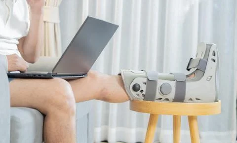 Side view shot of man with broken leg working on laptop at home sitting on so Stock Photos