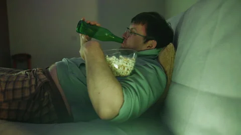 Side view of smiling man hugging bowl with popcorn and drinking from bottle Stock Footage 314464208