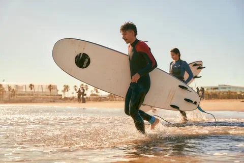 Side view of surfers in wetsuits running into the sea holding surfboards Stock Photos