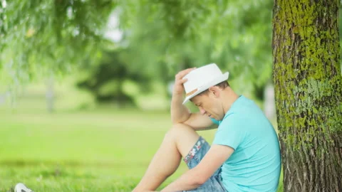 Side view of a tired man in a hat resting in the shade of a tree in the park Stock Footage 149531193