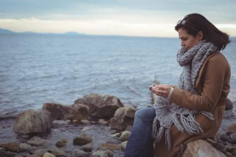 Side view of woman using mobile phone while sitting on driftwood 写真素材