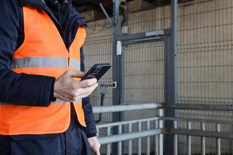 Side view of worker checking phone in reflective vest at fenced site Stockfoto's