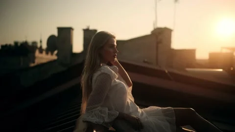 Side View of Young Attractive Blonde Woman in a white Dress Sitting on a Roof at Stock Footage 160158358