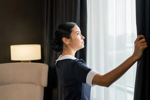 Side view of young chambermaid in uniform opening dark curtains on large window Stock Photos