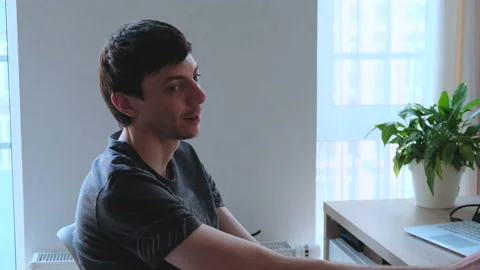 Side view of a young dark-haired man sitting at a table in front of a laptop 스톡 동영상 137105518