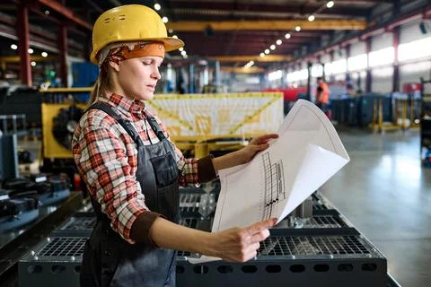 Side view of young female engineer in workwear holding unrolled blueprint Stock Photos