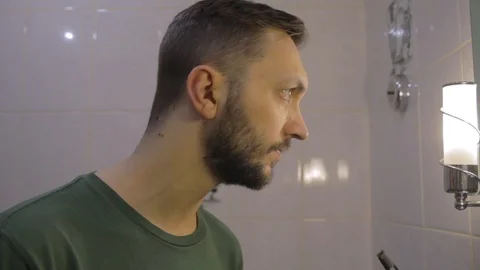 Side view on young man in bathroom looking at the mirror, checking face for skin Видео 104022012