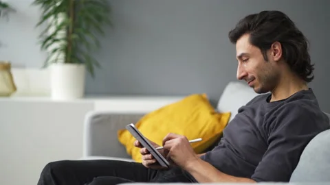 Side view of young man drawing with digital tablet while sitting on the sofa Stock Footage 149250994